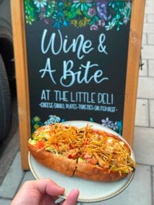 Loaded deli sandwich held before floral wine bar sign